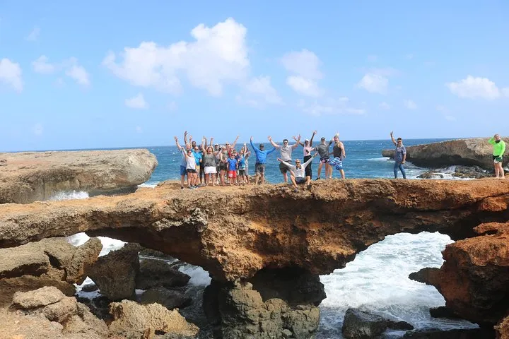Group of people standing on a rocky arch by the sea with arms raised.