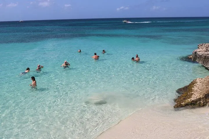 People swimming in clear turquoise ocean near a sandy beach with rocks.