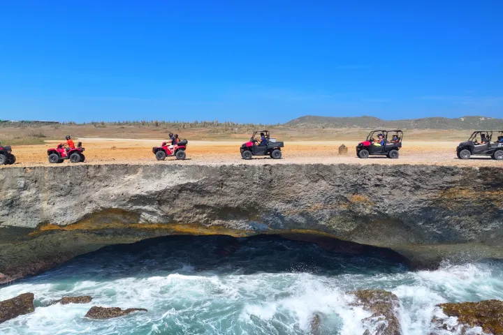 Five off-road vehicles on a cliff by the ocean under a clear blue sky.