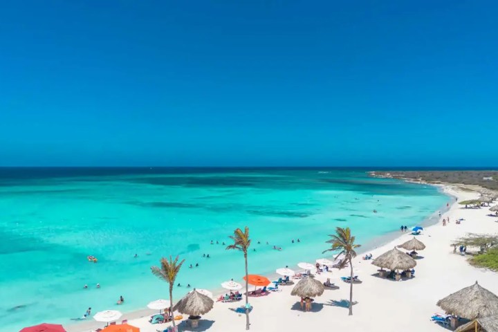 Tropical beach with turquoise water, palm trees, umbrellas, and people enjoying the sea under a clear blue sky.