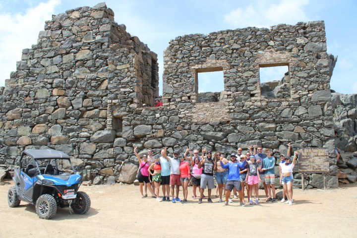 Group of people posing in front of old stone ruins with an ATV nearby on a sunny day.