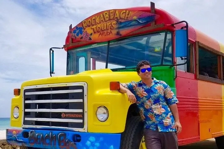 Man in floral shirt leaning on colorful tour bus with 'Rockabeach Tours Aruba' sign.