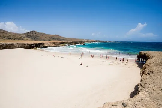 A sandy beach with people near the shoreline on a sunny day, rocky cliffs in the background.