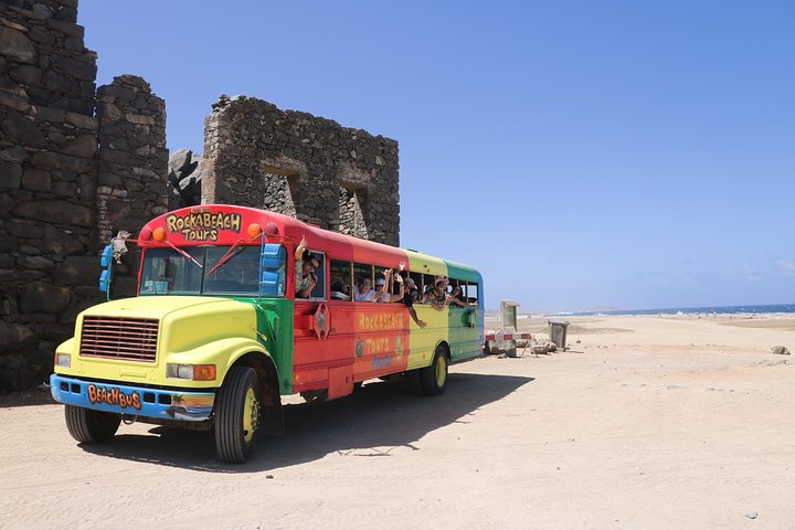 Colorful tour bus near a stone ruin on a sandy beach under a clear blue sky.