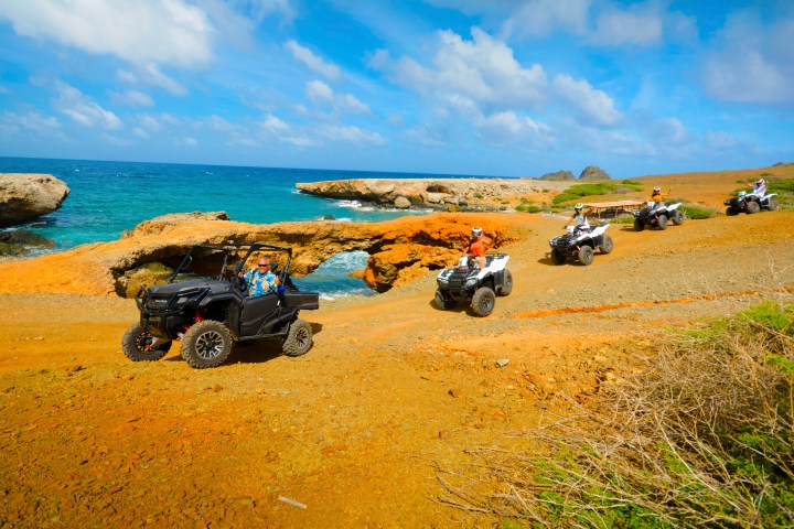 People on ATVs on a dirt path by the ocean with rocky arch and clear sky.