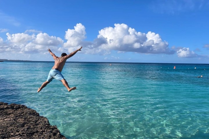 Person jumping off rocky cliff into clear blue sea under a bright sky with clouds.