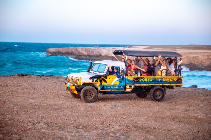 Open-top tour truck with waving passengers by a rocky coast and blue sea.