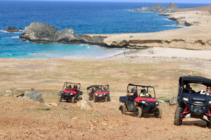 Four off-road vehicles driving up a rocky path near a coastline with blue ocean and sandy beach.
